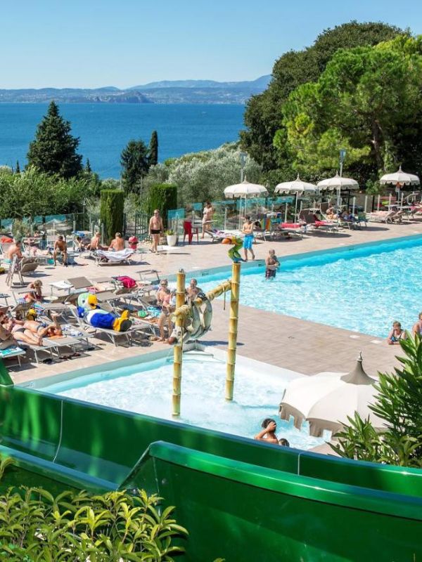 Lago di Garda Živá scéna u bazénu s lidmi opalujícími se a plavajícími. V popředí je zelená skluzavka a v pozadí klidný výhled na oceán.