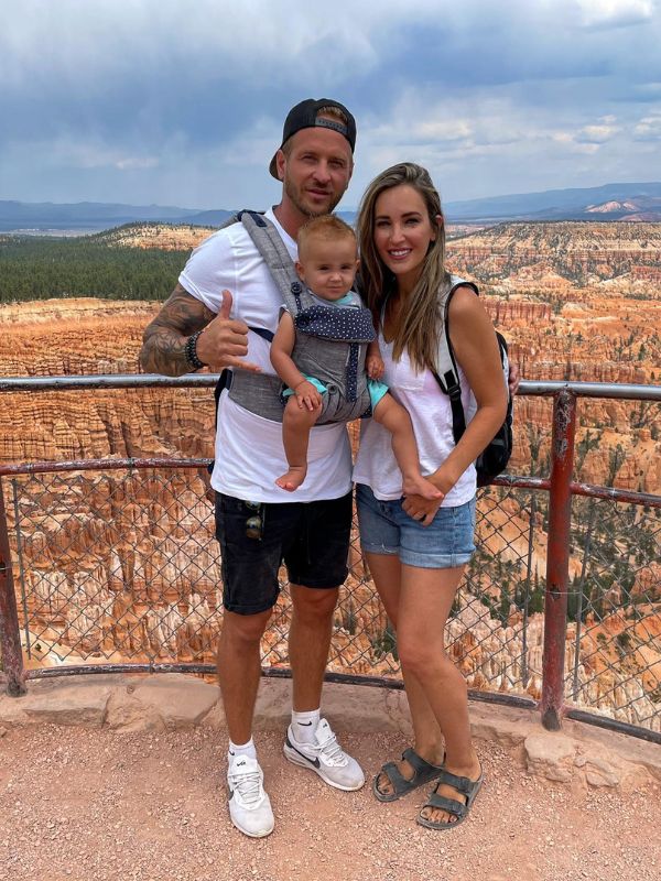 Bryce NP Usmívající se rodina pózuje na malebném vyhlídkovém místě s malým dítětem v nosítku. Za nimi se táhne úchvatná krajina kaňonu, která se rozprostírá do dálky.