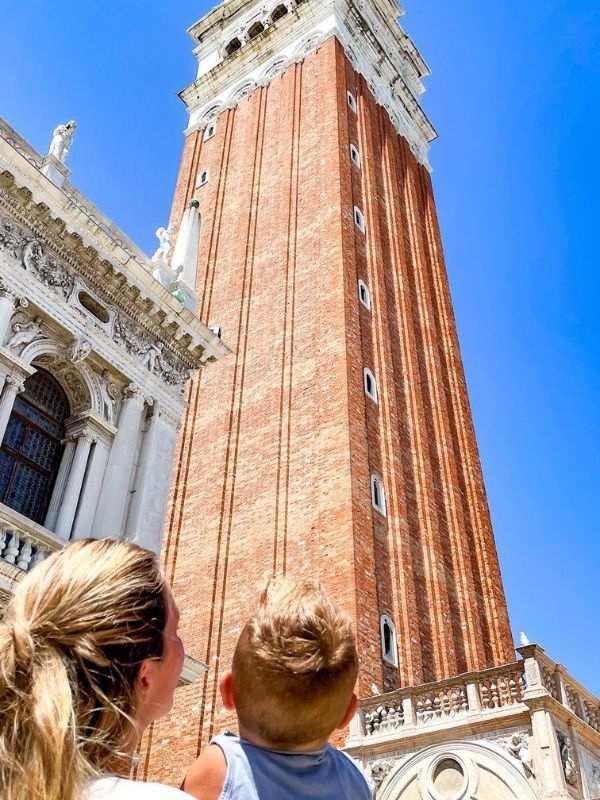 Campanile di San Marco Žena a dítě se dívají vzhůru na impozantní červenou cihlovou zvonici svatého Marka v Benátkách, proti jasně modré obloze, vyzařující údiv a zázrak.
