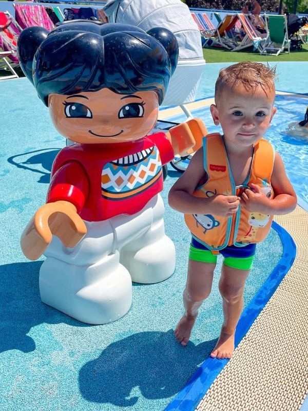 Lego Italy Mladé dítě v plavecké vestě stojí a usmívá se vedle velké sochy připomínající LEGO u bazénu. Jasný slunečný den s barevnými lehátky v pozadí.