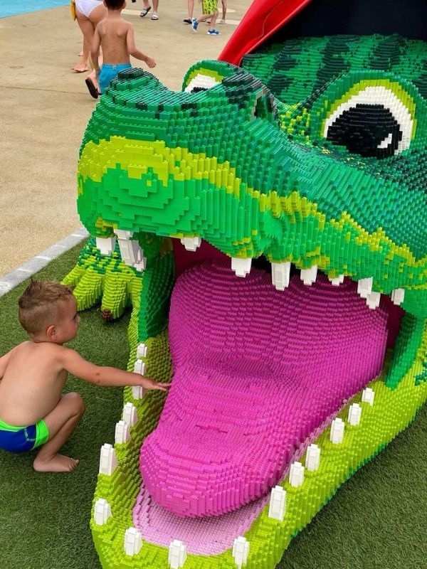 Lego Mladý chlapec v plavkách sahá na velký růžový jazyk hravé zelené LEGO sochy aligátora s otevřenou tlamou, což vyjadřuje zvědavost a zábavu.