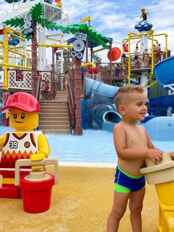 Legoland Mladý chlapec v plavkách se usmívá poblíž figur LEGO v životní velikosti na barevném vodním parku. Kolem něj jsou živé skluzavky, palmy a hravá atmosféra.
