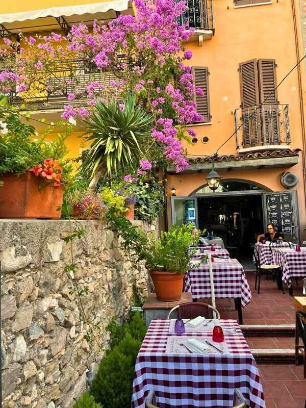Restaurace Venkovní italská kavárna s stolky pokrytými červenobílými kostkovanými ubrusy, obklopená kvetoucími růžovými květinami a bujnou zelení. Teplá a přívětivá atmosféra.