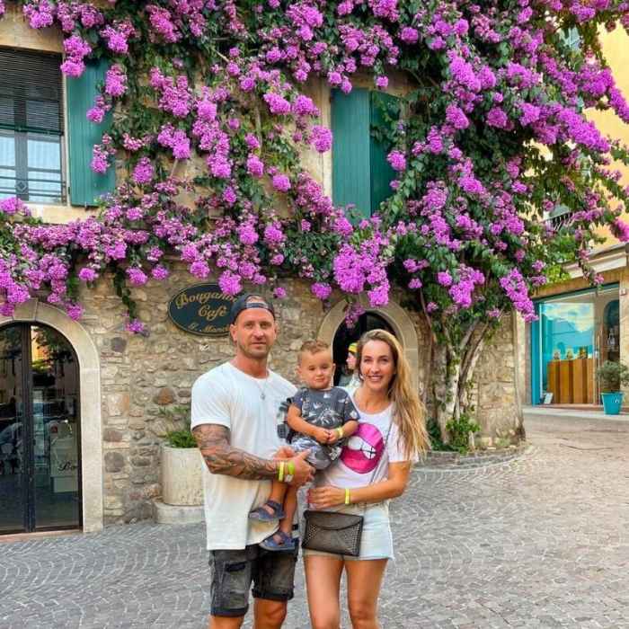 Rodina v Sirmione Rodina tří lidí se šťastně usmívá pod kvetoucími fialovými bugenvíliemi, které se sklánějí přes rustikální kamennou kavárnu s tyrkysovými okenicemi, čímž vytvářejí veselou a živou scénu.