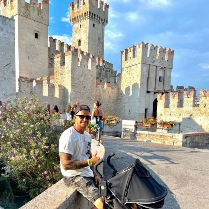 Sirmione Muž v bílém tričku a slunečních brýlích se usmívá poblíž kamenného hradu s cimbuřím. Sedí vedle kočárku a je obklopený kvetoucími rostlinami.