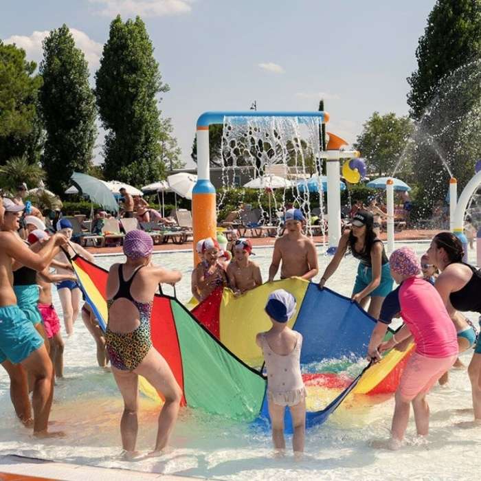 bazén hry Děti a dospělí si hrají v bazénu, zvedají barevný padák. Voda stříká a letní slunce vytváří radostnou, živou atmosféru.