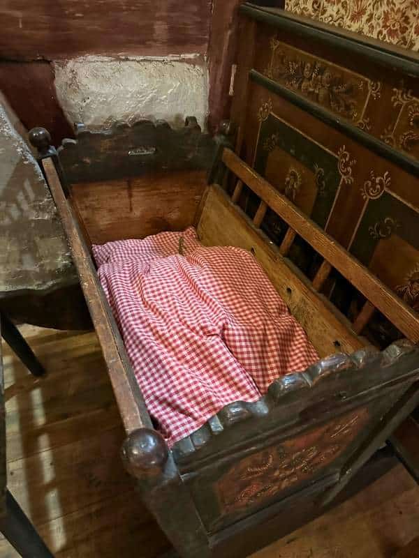 Baby Bed Seeberg Zdobená dřevěná kolébka s vyřezávanými detaily stojí v rustikální místnosti. Uvnitř je úhledně rozprostřená červenobílá kostkovaná deka, která navozuje útulnou, nostalgickou atmosféru.<br />