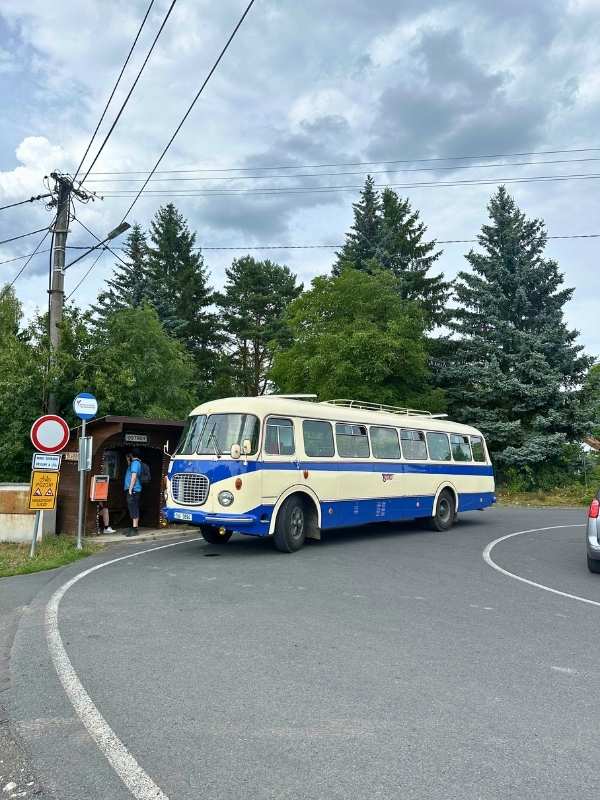 Bus Seeberg Retro modro-krémový autobus zatáčí na venkovské zastávce, v pozadí se tyčí vysoké zelené stromy. Zachmuřená obloha dodává scéně nostalgický nádech.<br />