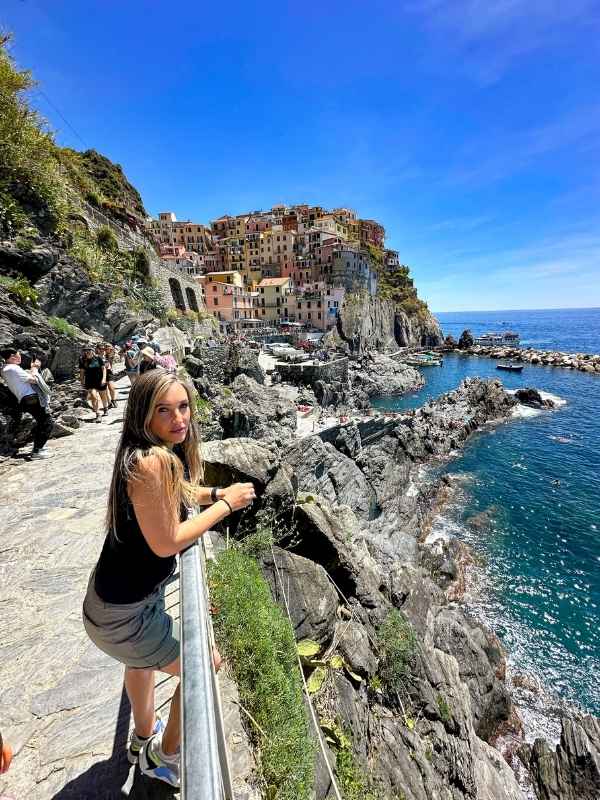 Manarola Žena stojící na skalnaté cestě s výhledem na živou přímořskou vesnici postavenou na útesu s barevnými domy. Scéna je jasná a živá, s průzračným modrým mořem a nebem.<br />
