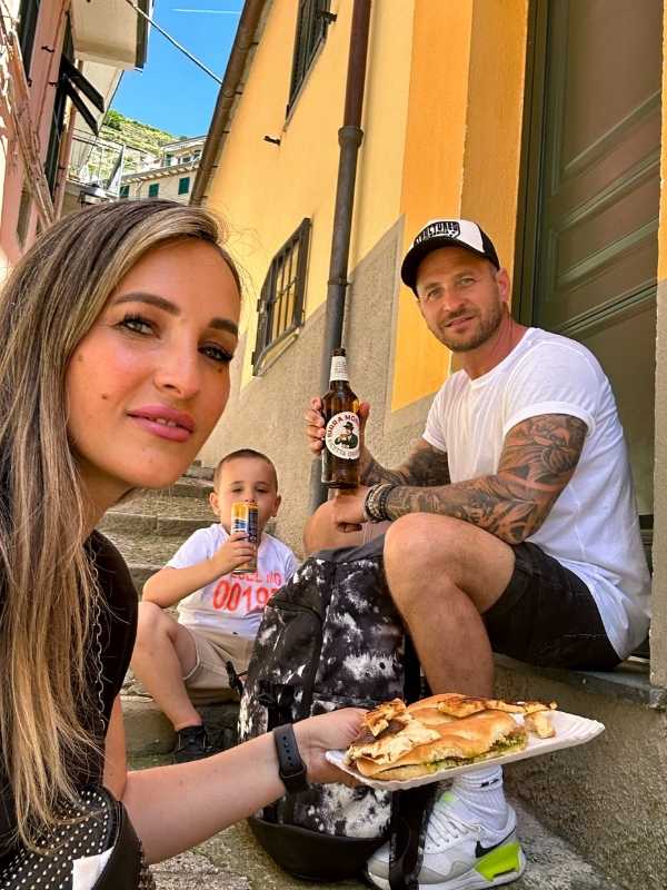 Manarola Cinque Terre Žena, muž a dítě sedí na venkovních schodech obklopeni pastelovými budovami. Žena drží sendvič, muž nápoj a dítě krabičku s džusem, všichni se usmívají.<br />