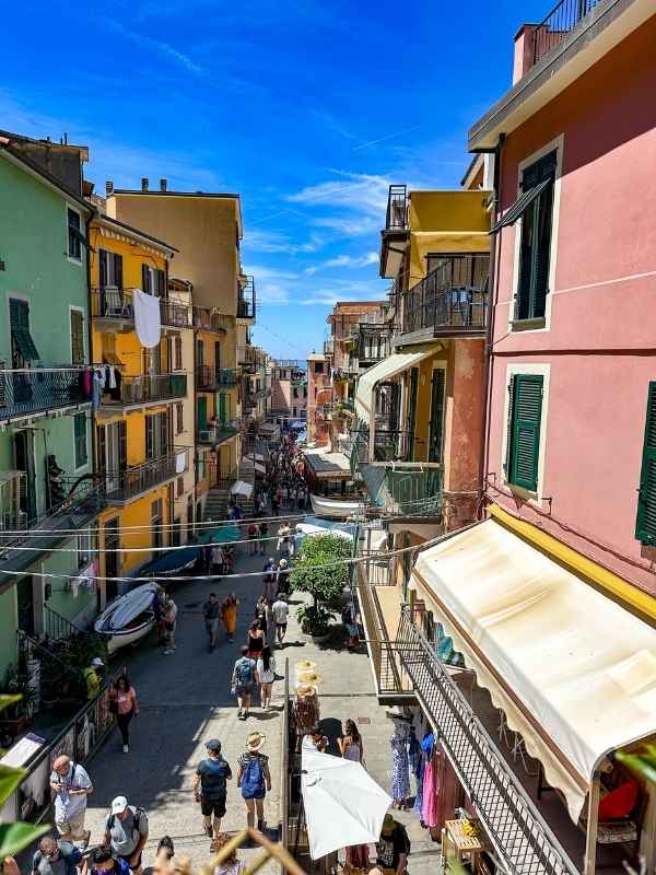 Manarola Italy Živá ulička v italské vesnici s barevnými domy a balkony. Turisté se procházejí pod jasně modrou oblohou, což vytváří živou atmosféru.<br />