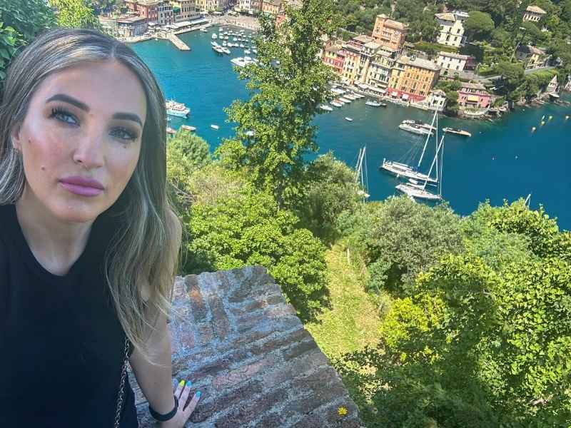 Portofino View Žena s dlouhými vlasy stojí u cihlové zdi a vyhlíží na živý přístav s plachetnicemi, barevnými budovami a bujnou zelení. Scéna působí uvolněně a malebně.<br />