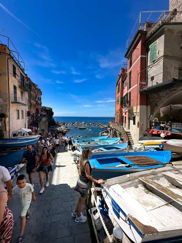 Riomaggiore Italy Úzká ulička lemovaná barevnými loďkami a živými budovami po obou stranách vede k skalnatému pobřeží a průzračnému modrému moři pod jasně modrou oblohou. Rušná atmosféra.<br />