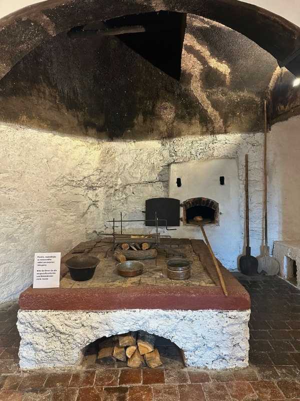 Seeberg Czech Historická rustikální kuchyně s černou klenutou střechou. Kamenná pec, dřevěné nástroje a hrnce vyzařují vintage a užitkovou atmosféru. Pod pecí je naskládané dřevo.<br />