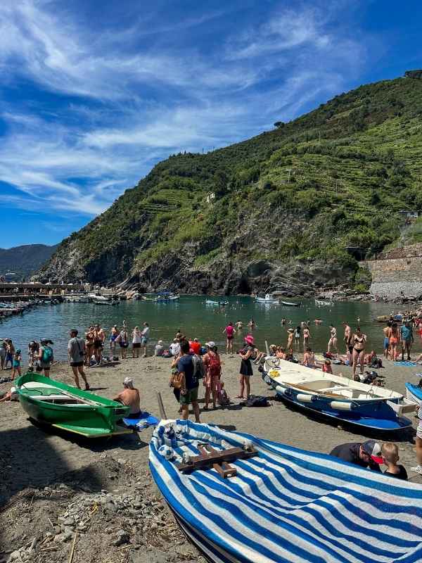 Vernazza Plážová scéna s lidmi odpočívajícími a plavci u zelených kopců a klidného moře. Barevné loďky jsou na písčitém břehu pod jasně modrou oblohou.<br />