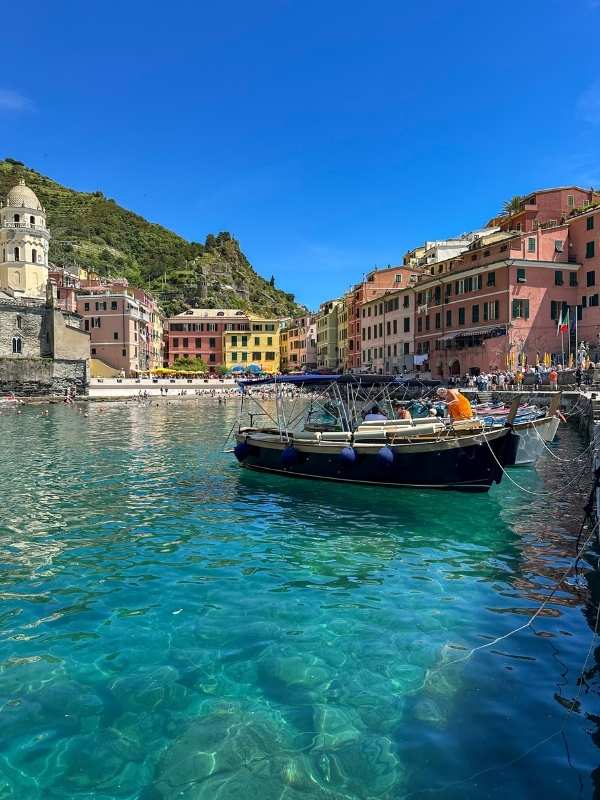 Vernazza - Cinque Terre Barevná přímořská vesnice s živými domy podél nábřeží. Průzračná modrá voda a slunečné nebe obklopují malou loď zakotvenou u břehu.<br />